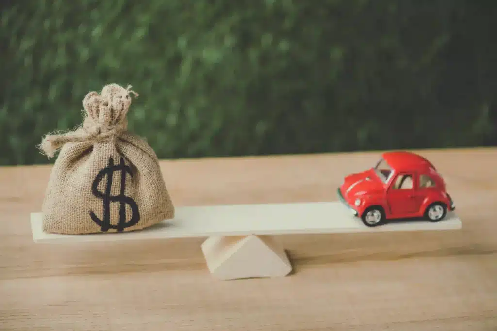 red miniature car and money bag with dollar sign balancing on seesaw on green background